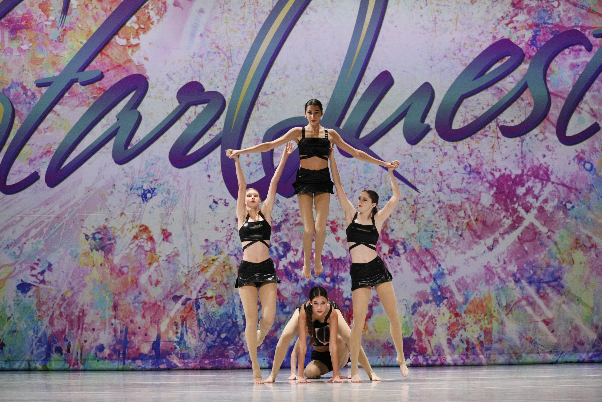 Five Zabava dancers in black costumes perform a synchronized lift and formation onstage at a youth dance competition.
