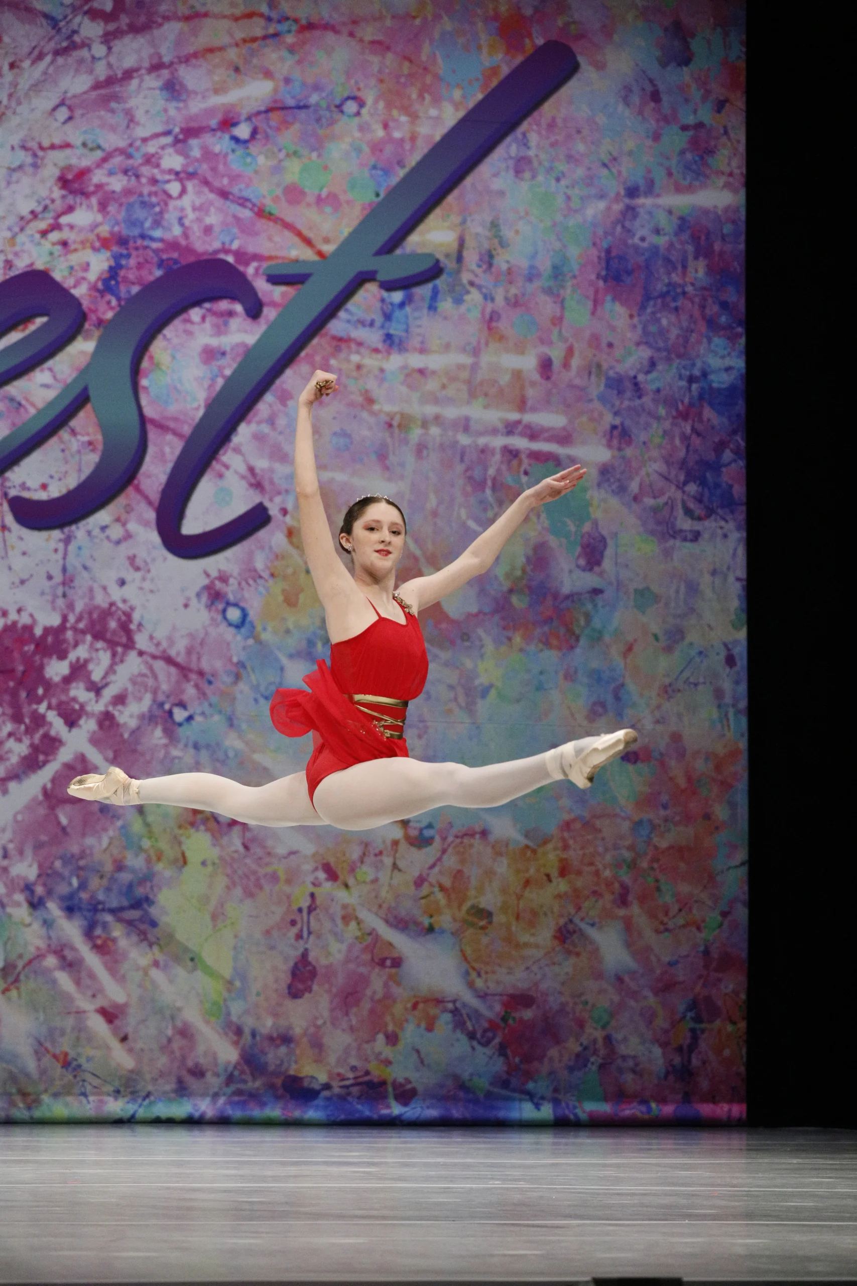 Zabava dancer in a red costume performs a split leap onstage during a youth dance competition.
